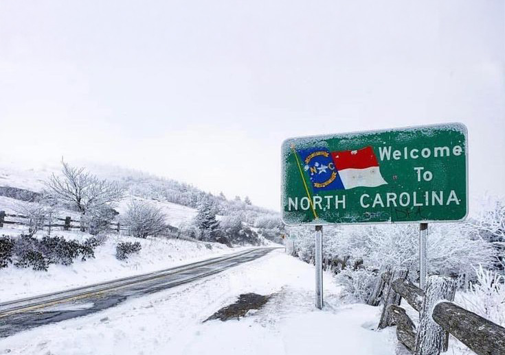 A snow-covered roadside and a frosted “Welcome to North Carolina” sign during a winter storm in the Great Smoky Mountains.